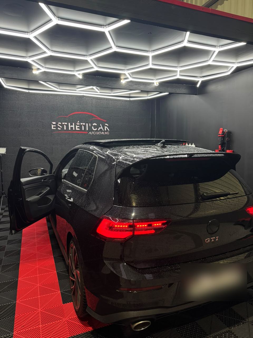 Golf GTI noire dans le box de l'atelier : plafond LED en nid d'abeille, logo Esthéti'Car au mur, sol damier rouge et noir